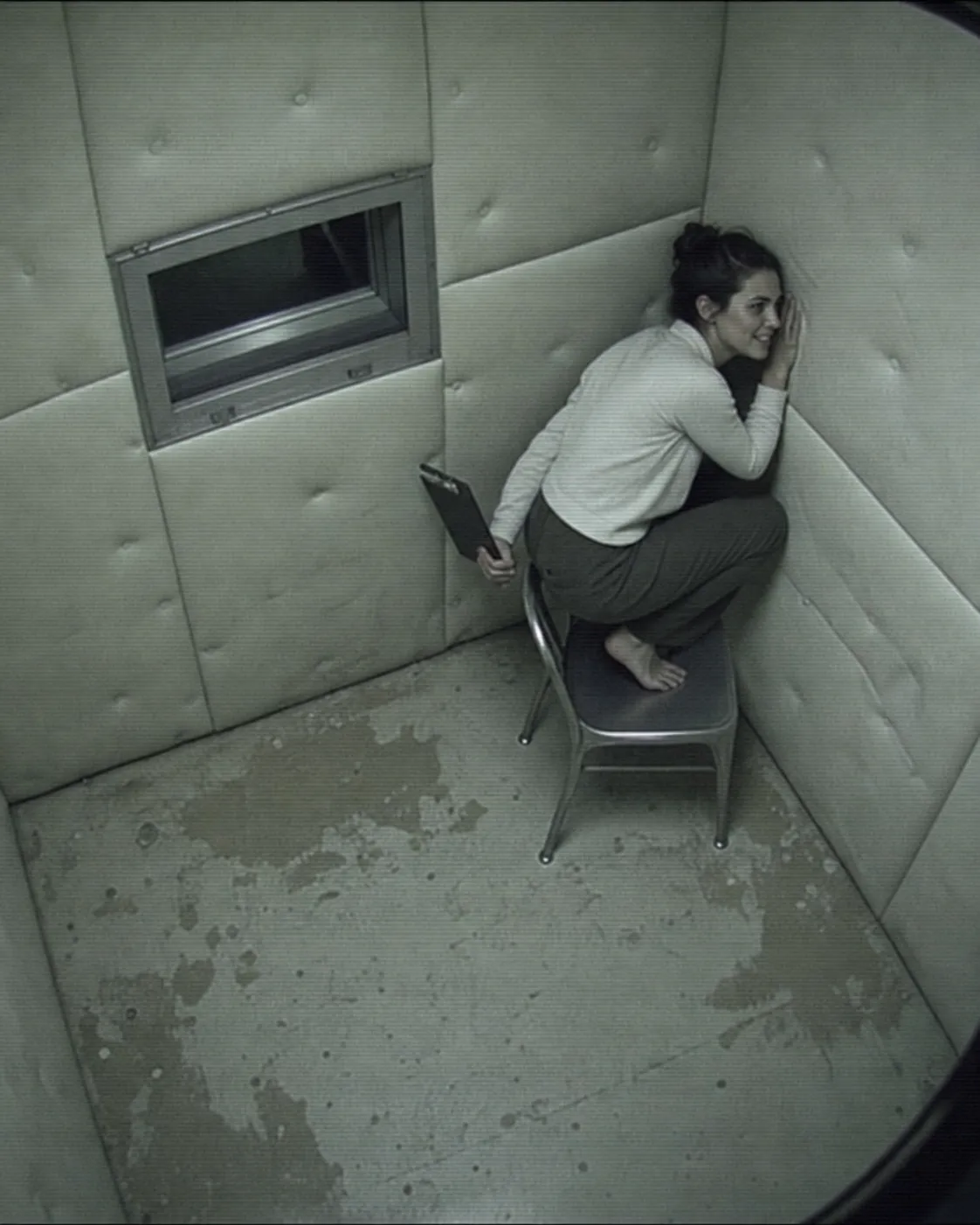 Security camera view of Mara Morrow listening at the wall of a padded observation room